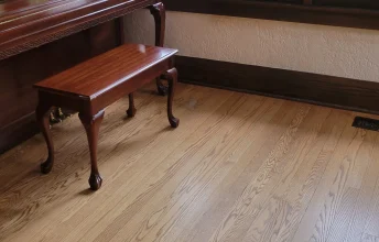 Shiny wooden piano next to dark brown wooden windows and a freshly mopped wooden floor.